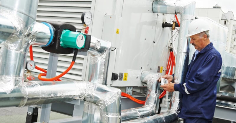 A worker in a blue uniform and white helmet inspects HVAC equipment with gauges and insulated pipes on a rooftop, ensuring peak performance for Chicago's diverse climate.