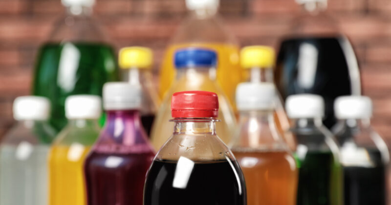 Assorted plastic bottles filled with various colorful beverages are displayed, featuring a focus on a bottle with a red cap in the foreground. These vibrant drinks, highlighted by best beverage distributor websites, stand out against a blurred brick wall background.