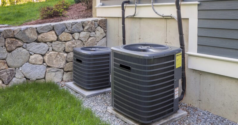 Two outdoor HVAC units rest on concrete slabs beside a house, framed by lush grass and a stone wall, ready to keep the indoors cool and comfortable.