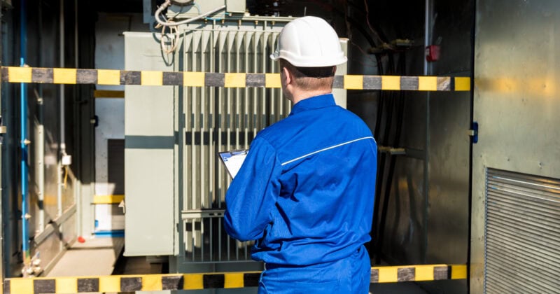 A person in blue protective clothing and a white hard hat inspects an electrical transformer, clipboard in hand, within a restricted area marked by yellow and black tape. Their work ensures seamless HVAC operations in this high-security zone.