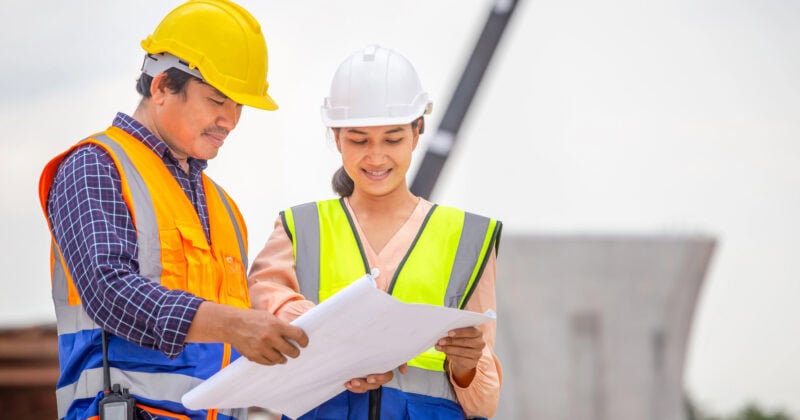 Two construction workers, clad in hard hats and safety vests, review blueprints at an outdoor site, discussing proven strategies that could double your leads in the industry.