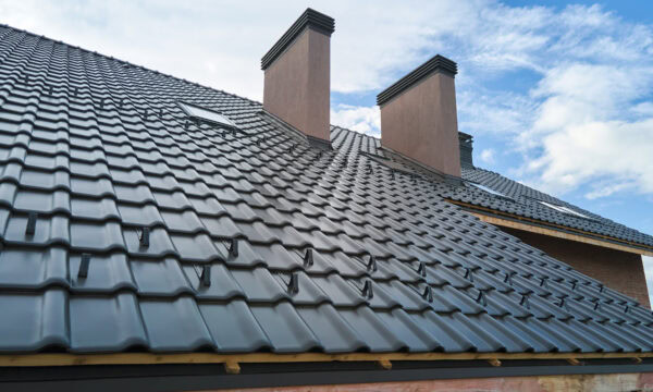 The image shows a house roof featuring black metal tiles and two brick chimneys against a partly cloudy sky, offering inspiration for roofing companies aiming to enhance their portfolios.