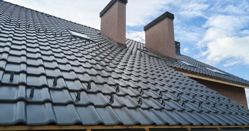 The image shows a house roof featuring black metal tiles and two brick chimneys against a partly cloudy sky, offering inspiration for roofing companies aiming to enhance their portfolios.