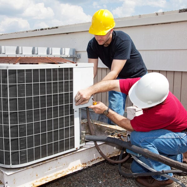 Two workers in safety helmets are repairing an outdoor air conditioning unit on a rooftop. One is standing, the other kneeling.