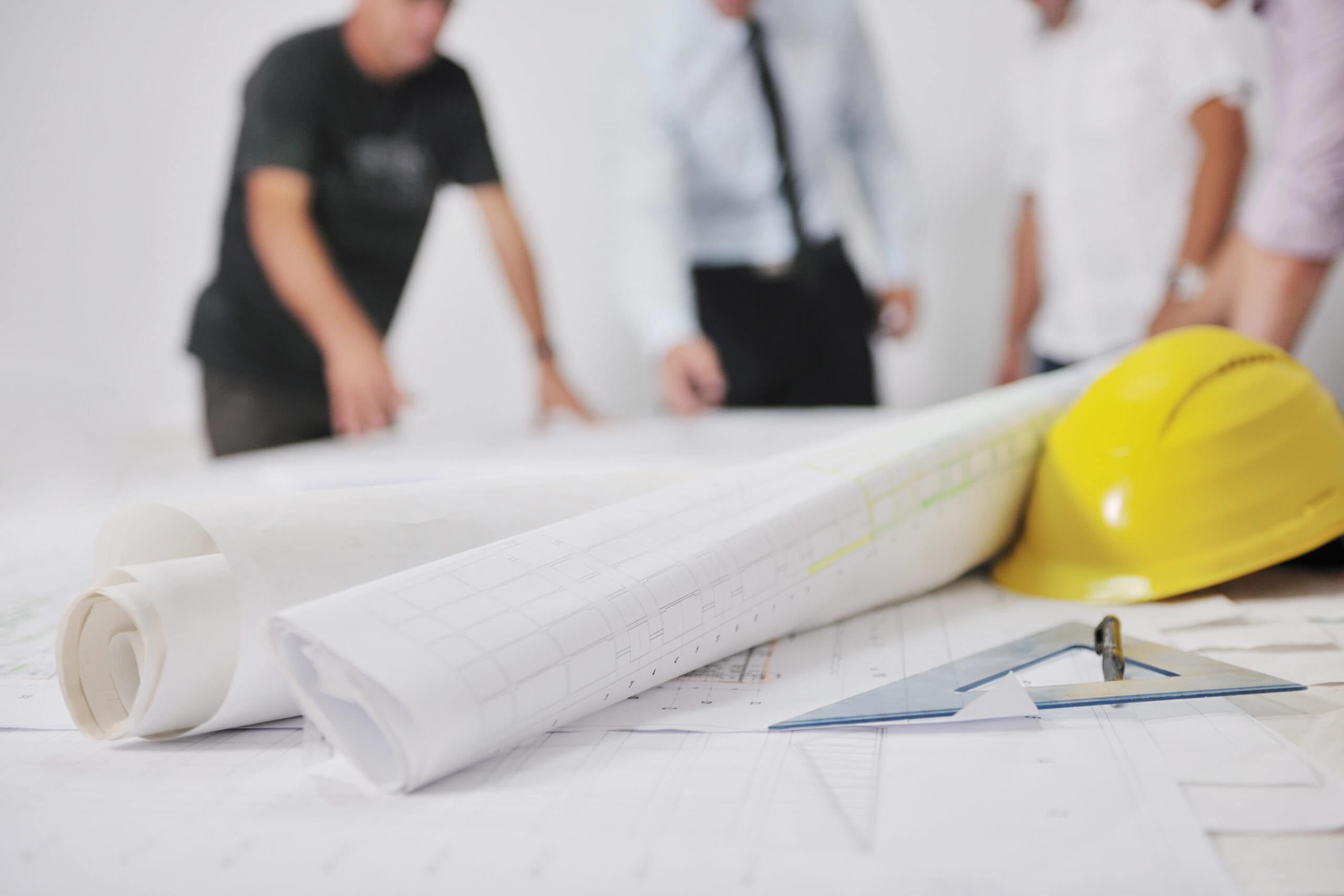 Blurred figures examine large architectural blueprints on a table. A yellow safety helmet, a pen, and a protractor are visible in the foreground.