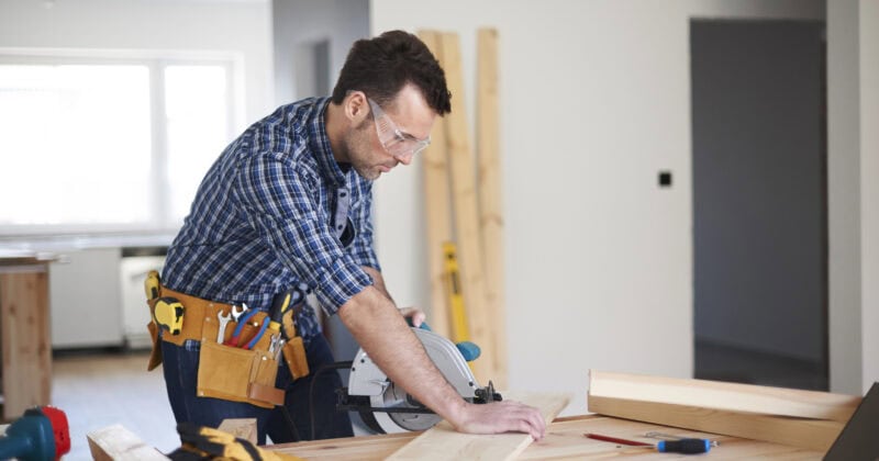 In a bustling workshop, a person wearing safety glasses and a tool belt skillfully uses a circular saw to convert wood into precise pieces, readying them for future contracting projects.