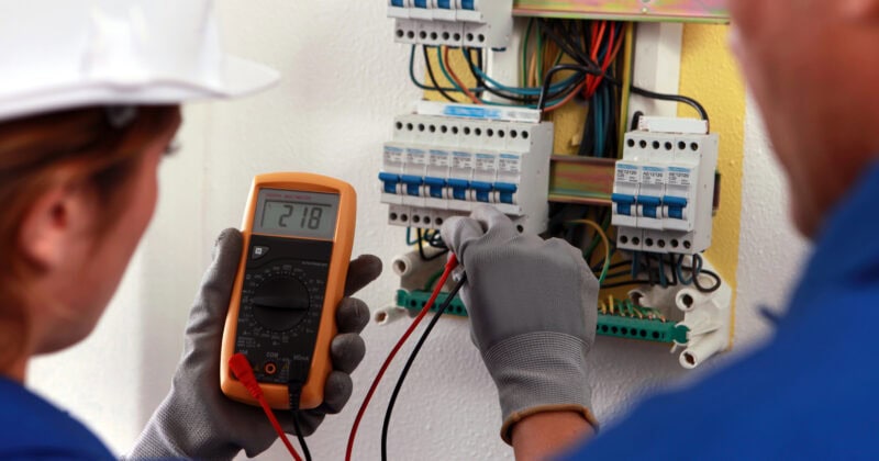 Two electricians wearing helmets and gloves work on an electrical panel; one holds a multimeter showing a reading of 218, illustrating a scene from the "2025 Blueprint" for Digital Success in Electrician Marketing.