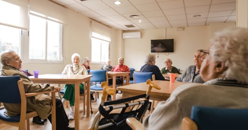 A group of elderly people sitting at tables in a well-lit room with large windows, a television on the wall, and a walker in the foreground. This is one of the best aspects of assisted living: creating vibrant communities that feel like home.