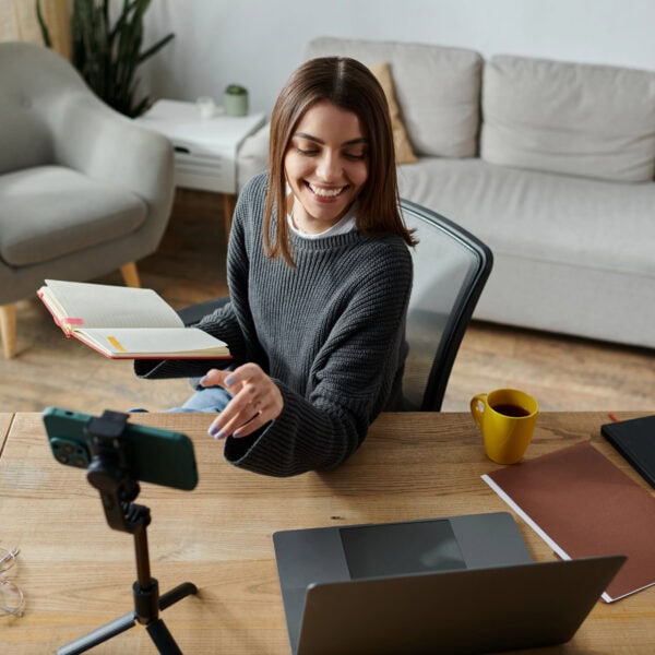 A woman sits at her desk, holding an open book and smiling at a smartphone on a tripod, likely recording something for her digital marketing agency. Nearby, a laptop, notebooks, and a cheerful yellow mug complete the scene.