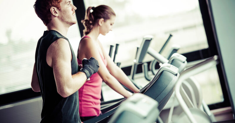 Two people exercise on treadmills in a gym. The man in the foreground wears a sleeveless shirt and gloves, while the woman behind him wears a pink tank top. Large windows are in the background, providing inspiration akin to top fitness websites where motivation thrives.