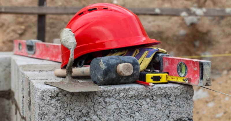 Construction tools rest on a concrete block: a red hard hat, spirit level, hammer, gloves, measuring tape, pencil, and trowel. This scene is a testament to the do's and don'ts of home services marketing—always ensure you have the right tools for the job.