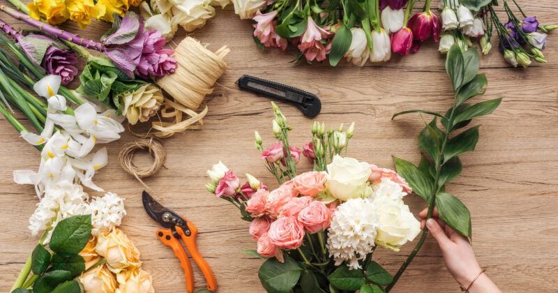 Hands arranging a floral bouquet on a wooden surface, surrounded by scattered flowers, ribbon, scissors, and a utility knife—much like scenes of creativity found on the best florist websites.