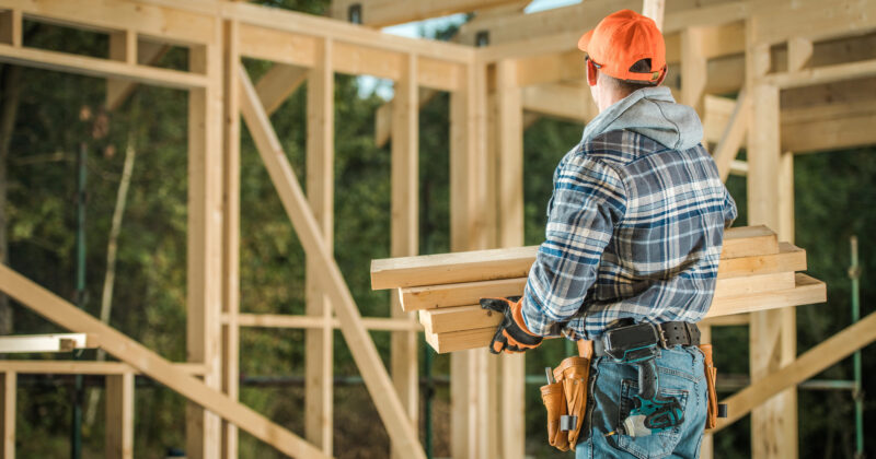 A construction worker in a plaid shirt and orange hat carries wooden planks at a wooden frame structure site, embodying the craftsmanship highlighted on the best home builder website design platforms.