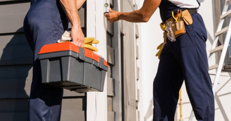 Two workers in blue uniforms are repairing a building. One holds a toolbox, while the other uses tools on a door—a scene reminiscent of optimizing your website for seamless lead conversion.