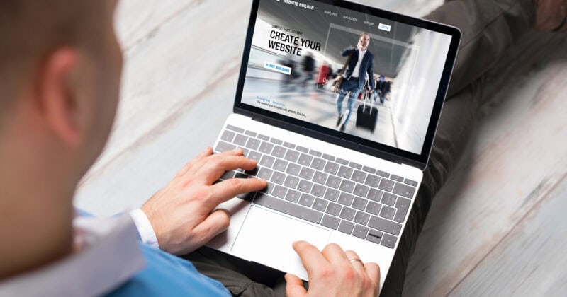 A person sits with a laptop on their knees, viewing a webpage that says "Create Your Website." The screen displays a workspace interface, perfect for business owners exploring platforms like WordPress.