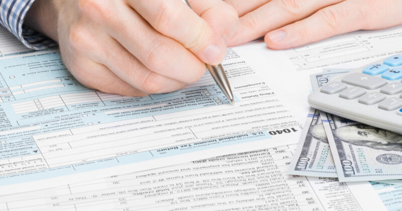 A person diligently fills out tax forms with a pen, surrounded by a calculator, currency, and a laptop open to their favorite CPA websites for guidance.