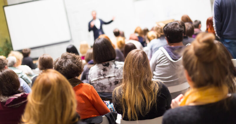 A speaker addresses a seated audience in a brightly lit room at the best conference. Participants are focused on the presentation, with a large screen visible in the background, showcasing content from top conference websites.