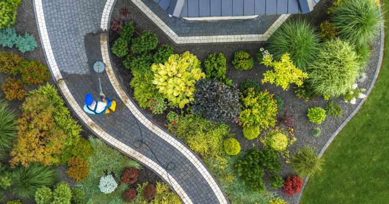 Aerial view of a landscaped garden with a stone path. A person in blue and yellow attire is watering plants on the curved walkway, surrounded by various shrubs and greenery.