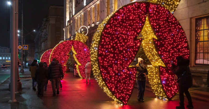 People stroll past large, illuminated red ornaments adorned with yellow tree designs on a city street at night, marveling at the captivating Holiday Light Installation.