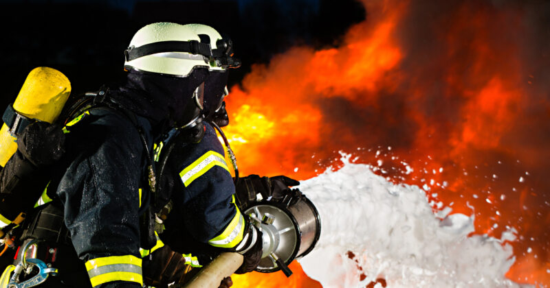 Firefighters in protective gear, featured on Fire Department websites, spray foam to extinguish a large fire, set against bright orange flames in the background.