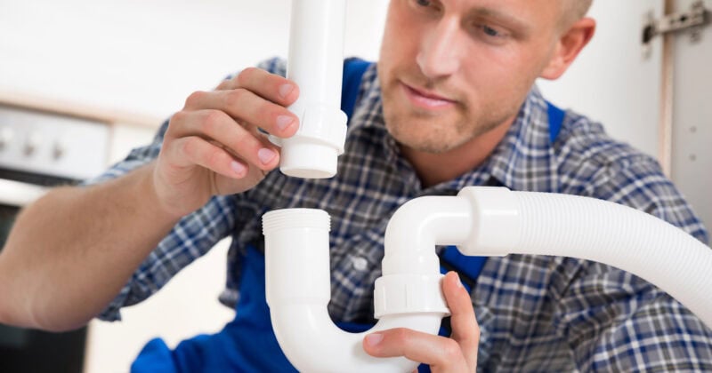 A person in a plaid shirt and blue overalls is assembling white plastic plumbing pipes under a sink.