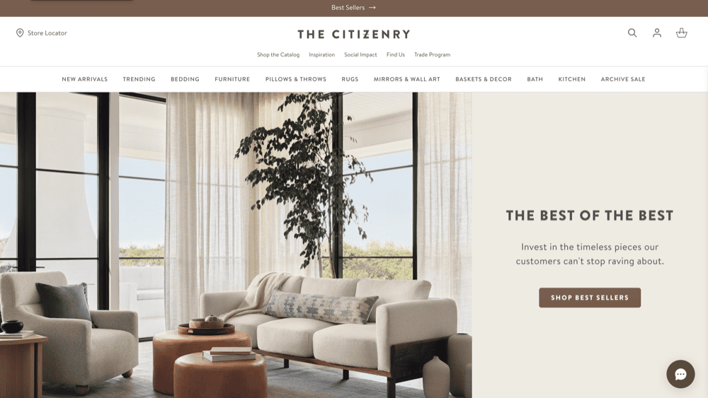 Experience modern elegance in this living room featuring a light gray sofa, lush tree, and cozy brown poufs. With large windows framed by curtains, it's a sneak peek into 2025's best home decor. Discover "The Best of the Best" from The Citizenry on top home decor websites today.